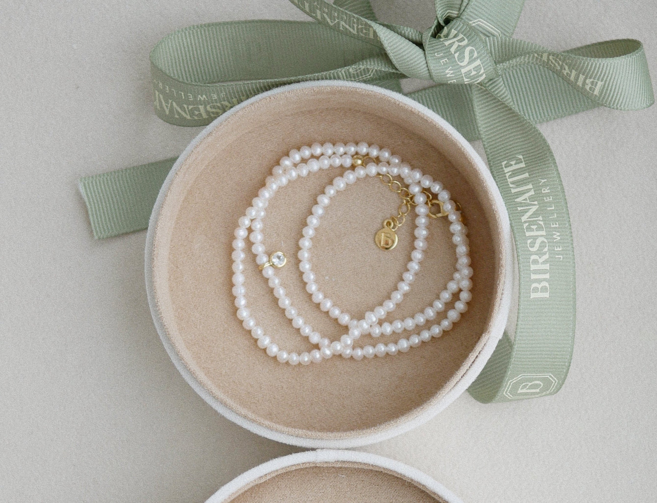 Open jewelry box with pearls and a gold necklace, branded 'Birsenate', on a light background.