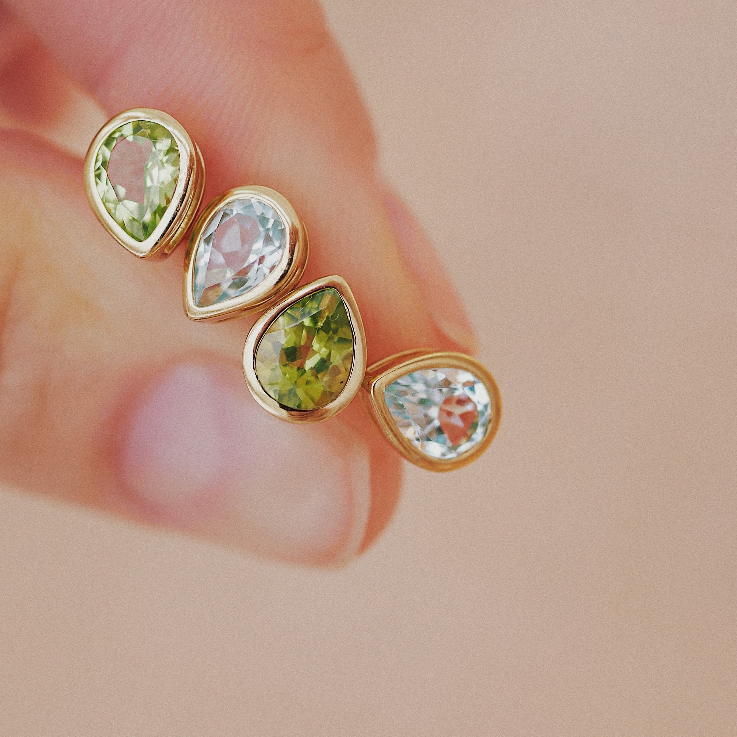 Gold earrings with green and clear gemstones held between fingers on a beige background