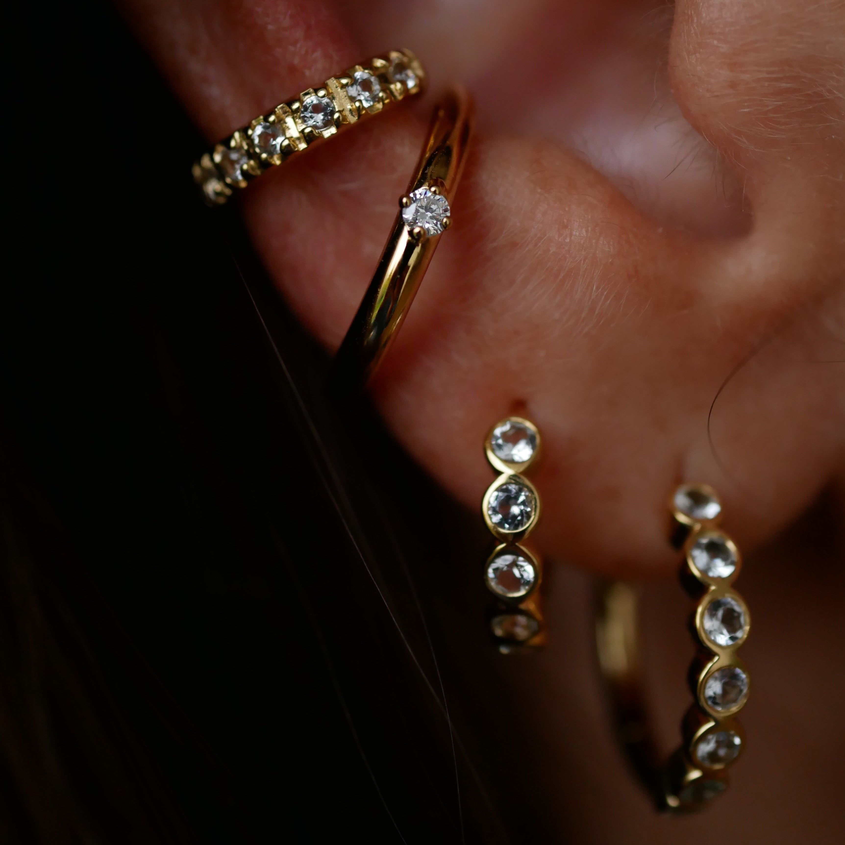 Gold hoop earrings with clear gemstones on an ear against a dark background