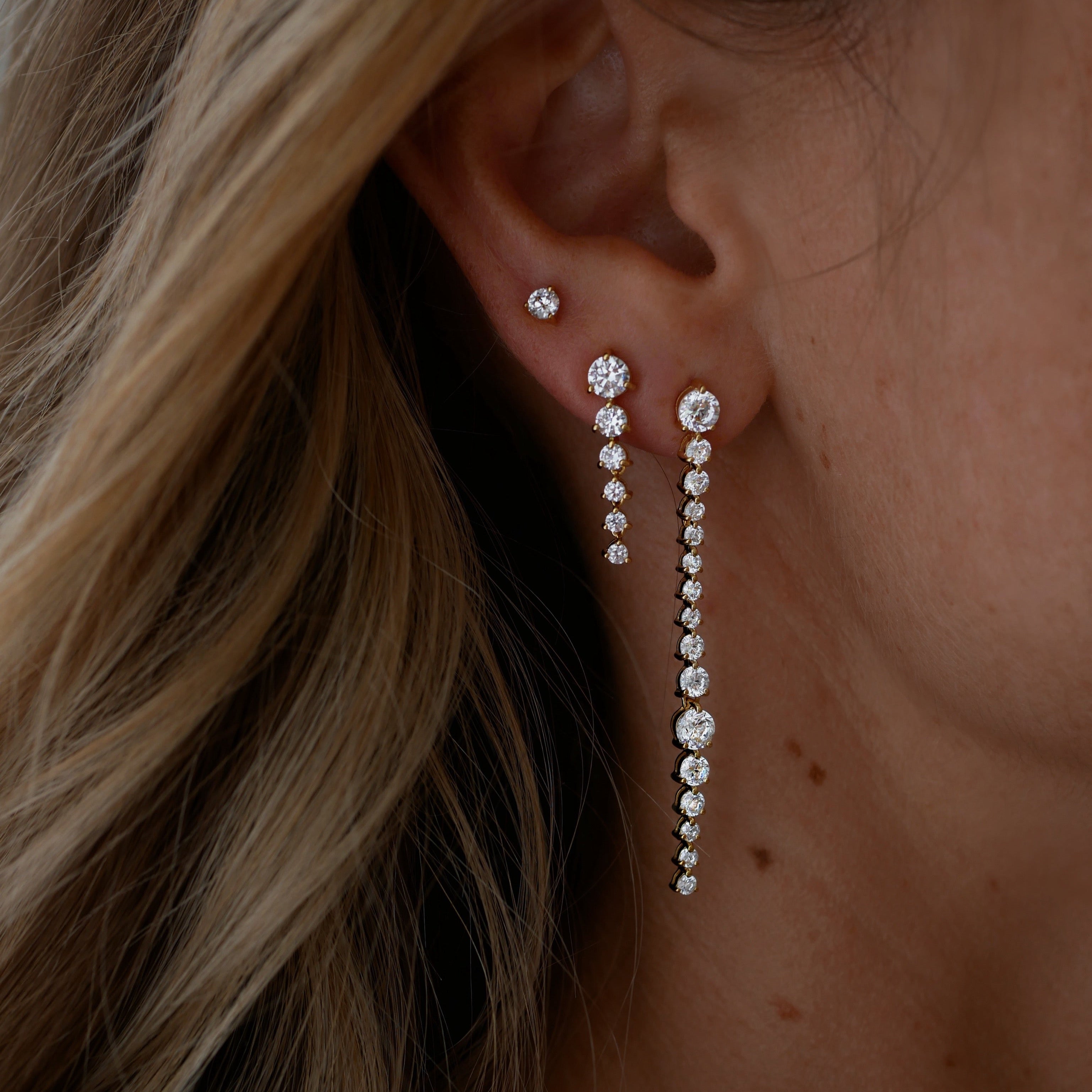 Close-up of an ear wearing a pair of dangling cubic zirconia earrings with blonde hair partially covering the face.