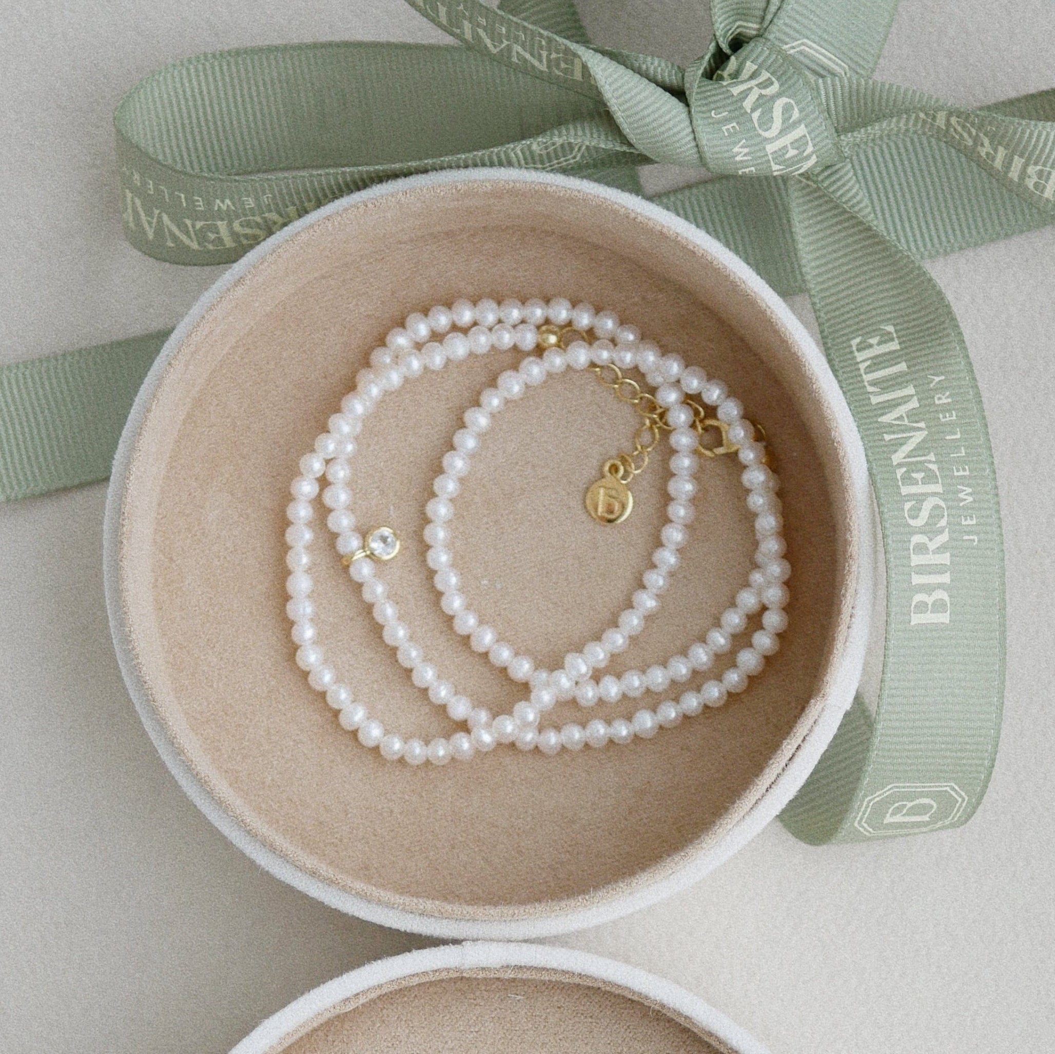 Open jewelry box with pearls and a gold necklace, branded 'Birsenate', on a light background.