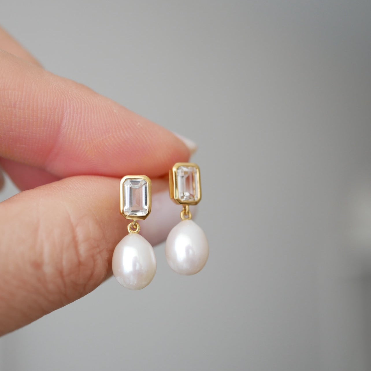 Pearl earrings with gold accents held by a hand against a gray background