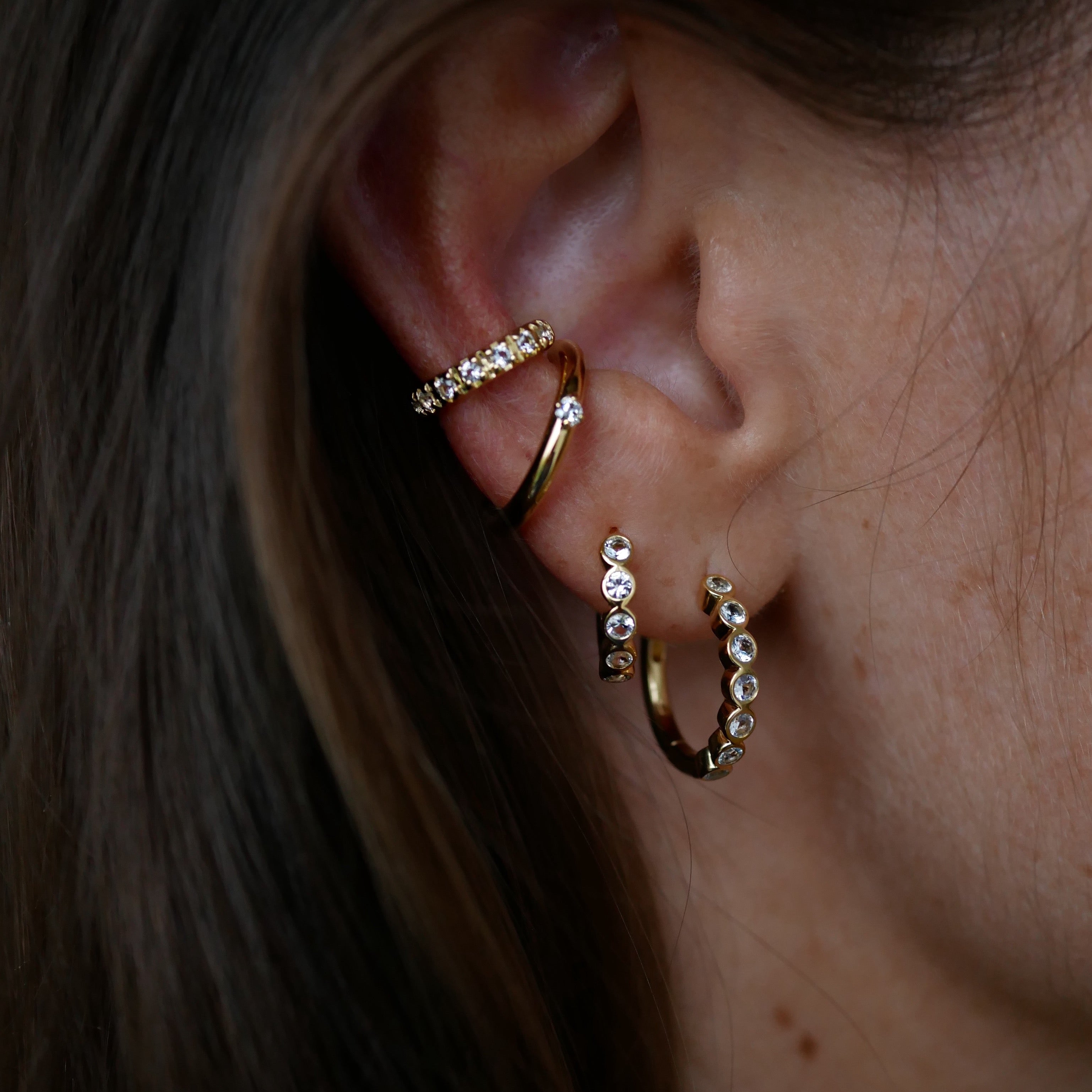 Close-up of an ear wearing gold hoop earrings with small stones.